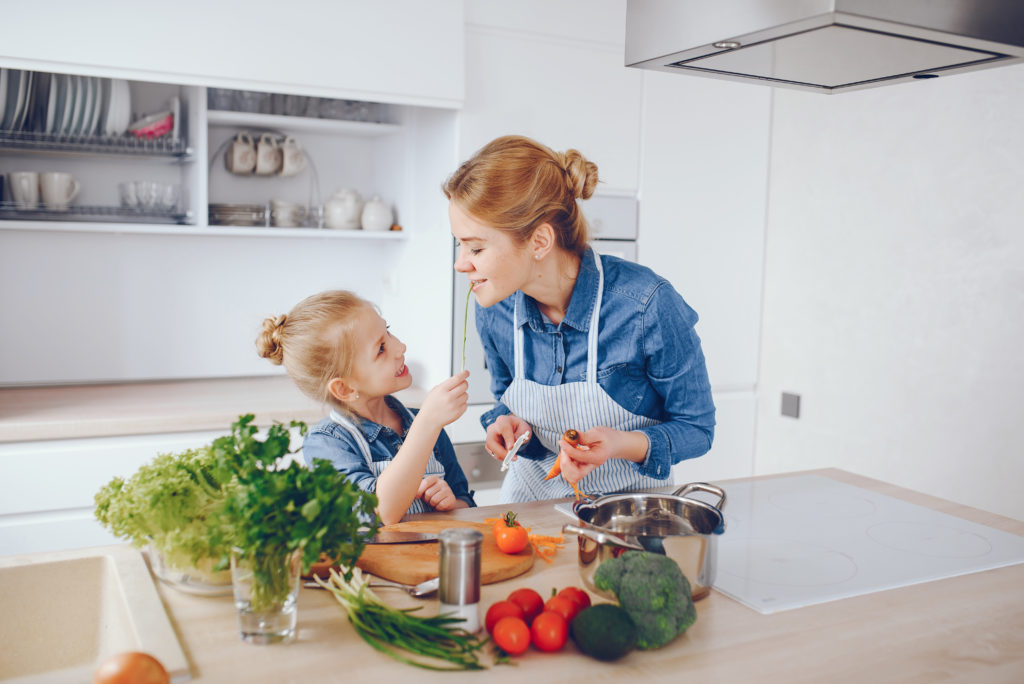 A young and beautiful mother in a blue shirt and apron is preparing a fresh vegetable salad at home in the kitchen, along with her little cute daughters with light hair