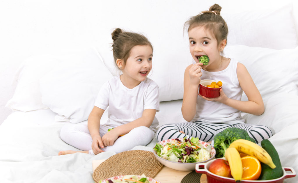 Healthy food at home. Happy two cute children eating fruits and vegetables in the bedroom on the bed. Healthy food for children and teenagers.